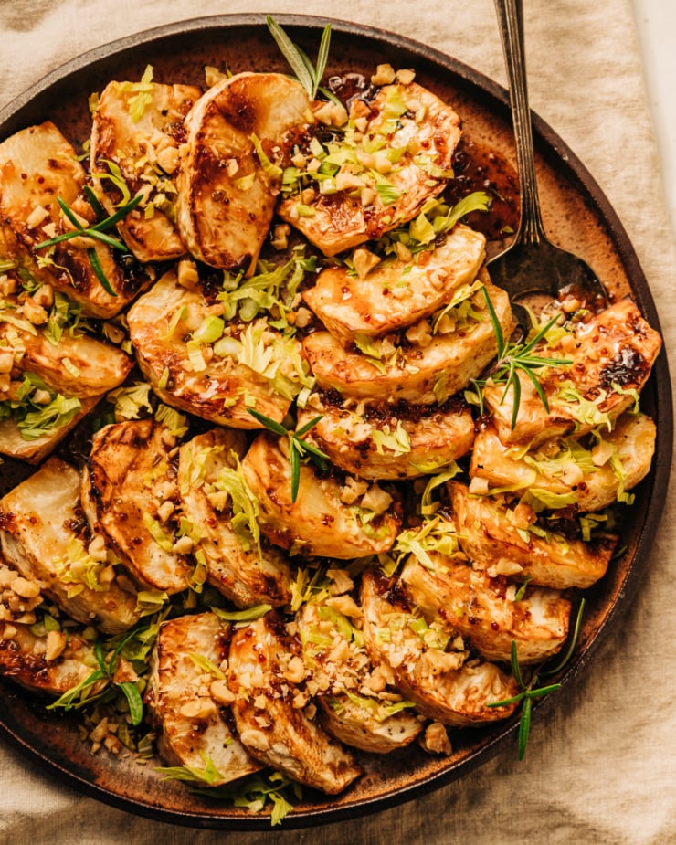 An overhead shot of cider glazed celery root pieces fanned out on a speckled brown plate. The celery root is topped with chopped walnuts, rosemary leaves, and finely chopped celery heart leaves.