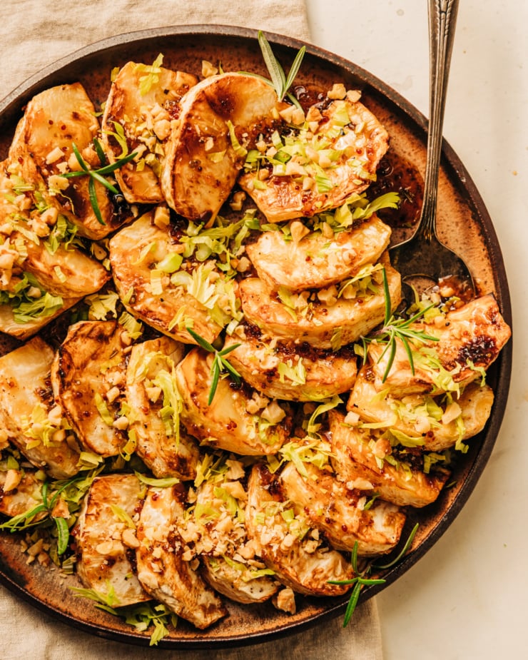 An overhead shot of cider glazed celery root pieces fanned out on a speckled brown plate. The celery root is topped with chopped walnuts, rosemary leaves, and finely chopped celery heart leaves.