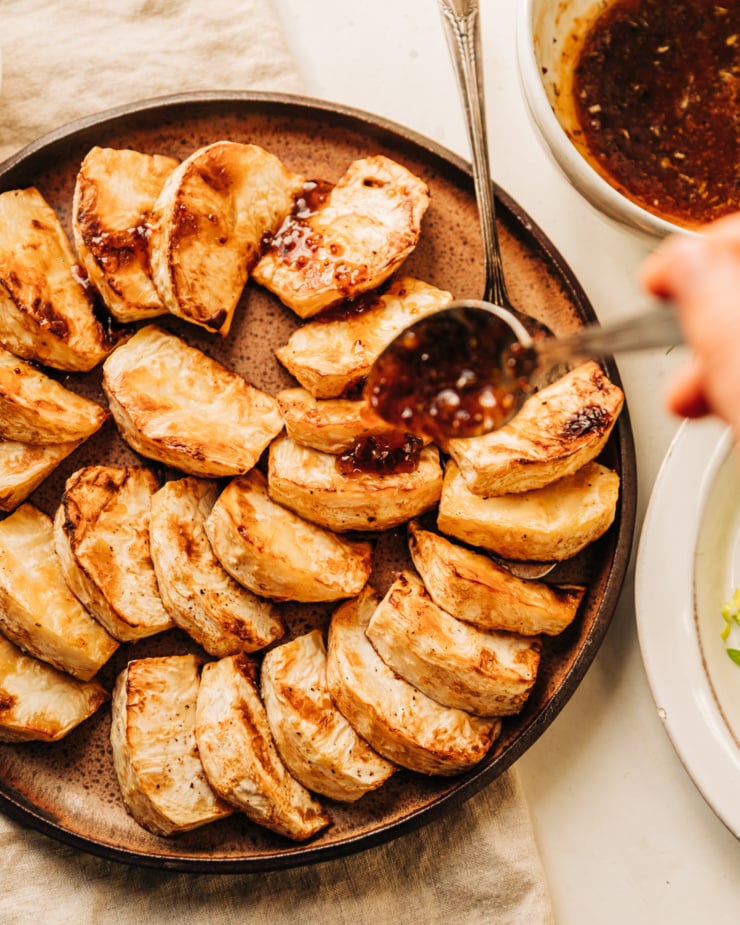 An overhead shot of roasted celery root pieces fanned out on a plate. A hand is using a spoon to glaze the pieces with apple cider reduction.