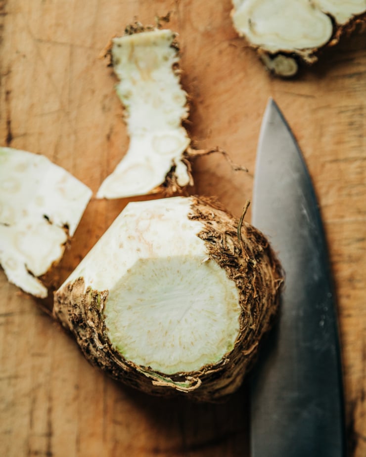 An overhead shot shows the process of a celery root being peeled with a chef's knife.