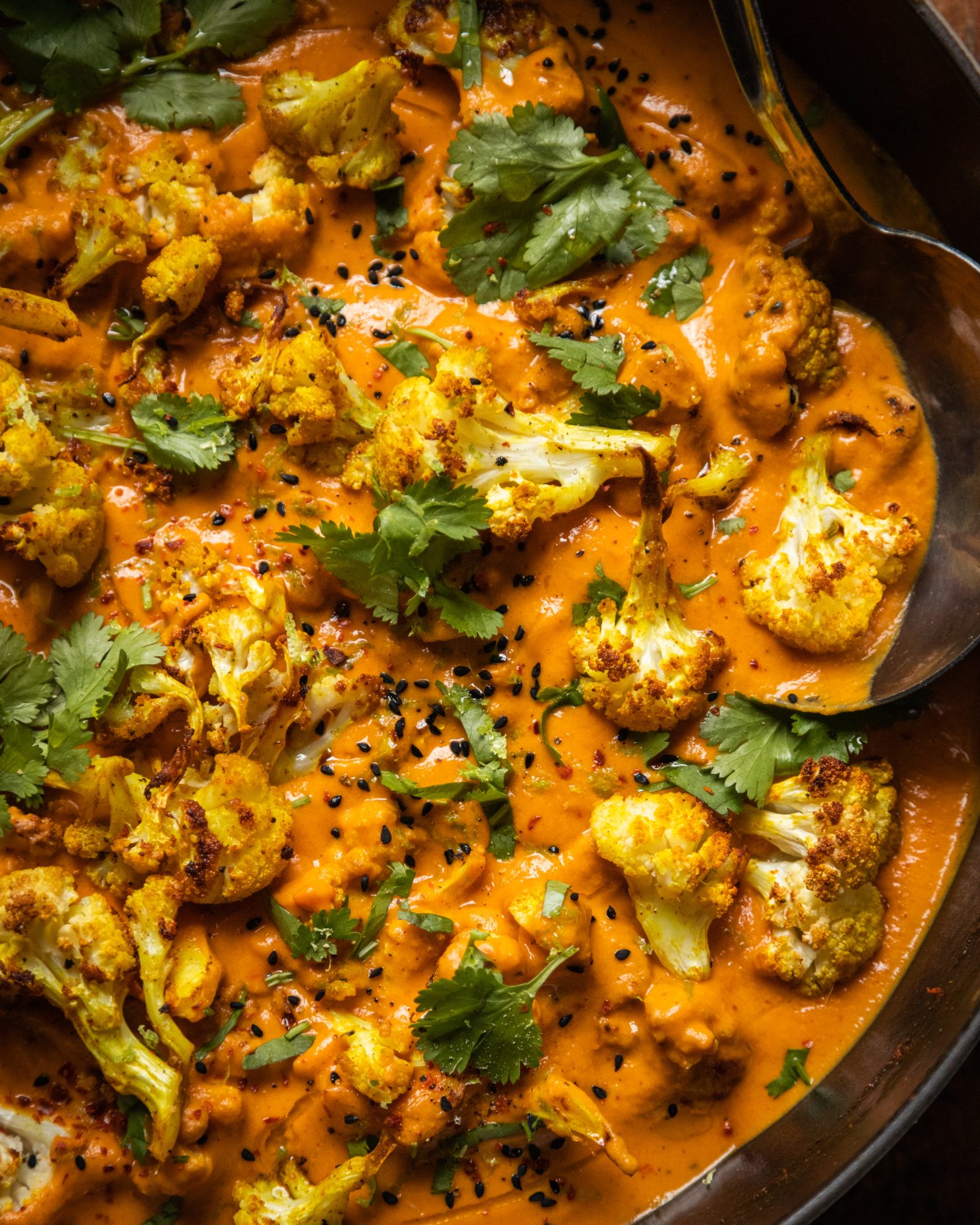 An up close, overhead shot of roasted cauliflower in a coconut tomato sauce. The dish is garnished with fresh cilantro, nigella seeds, and ground chillies.