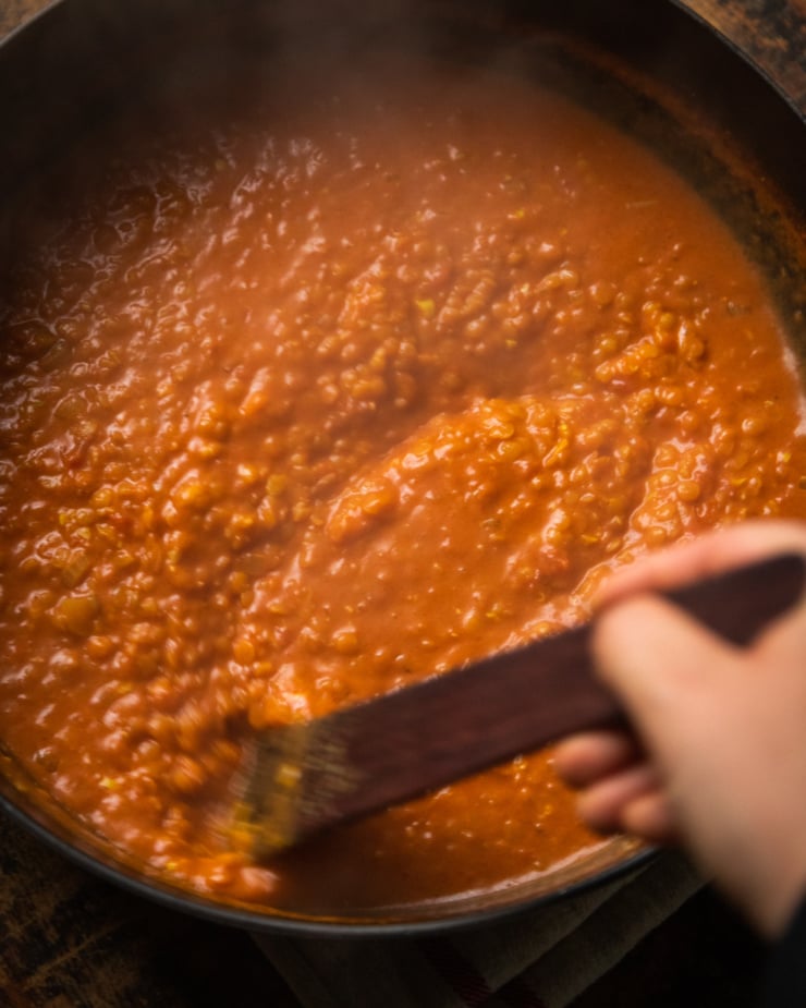 An overhead shot of a hand using a wooden utensil to stir up a pot of coconut tomato sauce with red split lentils.