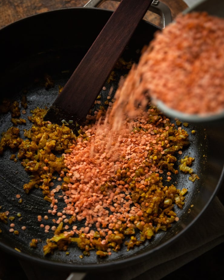 A slight 3/4 angle shot shows split red lentils being added to a pot of sautéed shallots and spices.