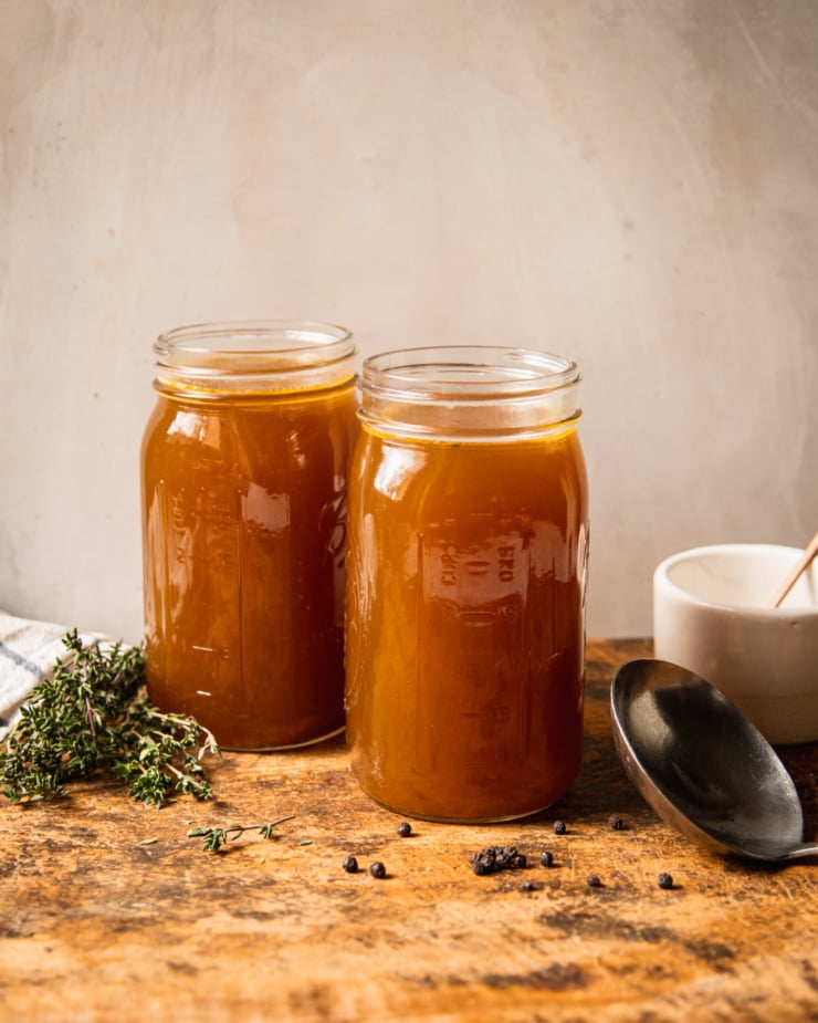 A head-on shot shows two jars of vegetable stock on a rough wooden cutting board. Sprigs of thyme, black peppercorns, a ladle, and a pot of salt are seen to the side.