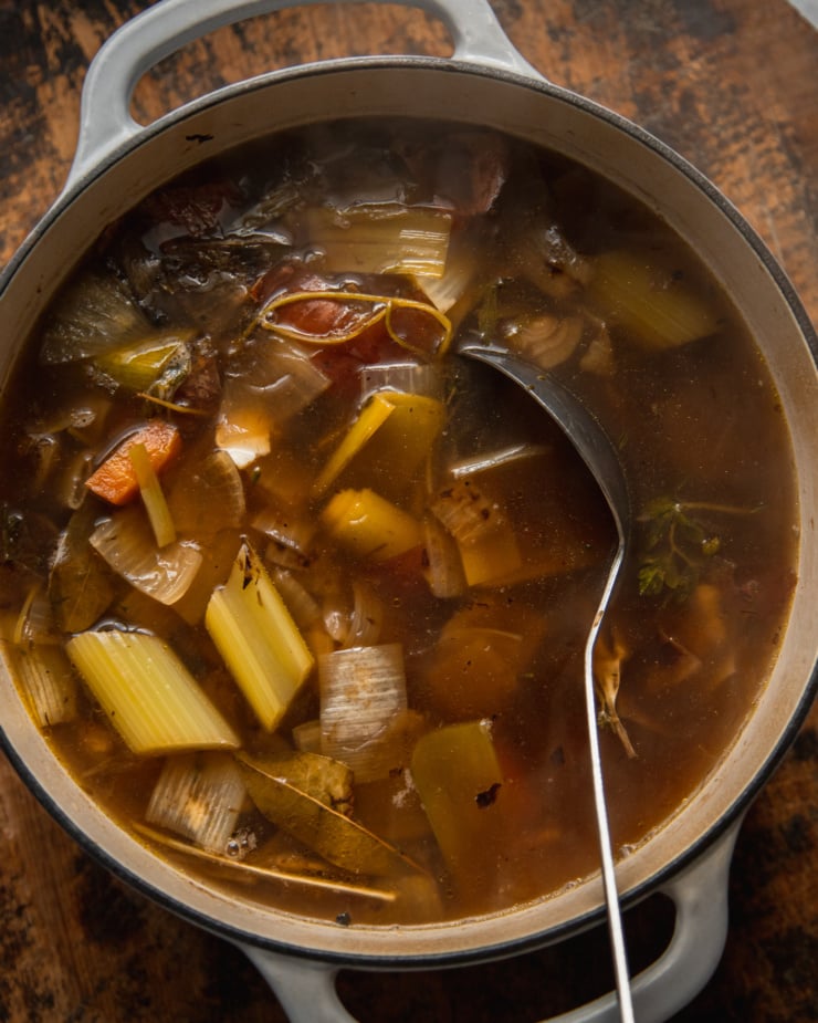 An overhead shot shows a finished pot of vegetable stock with the cooked vegetables and herbs yet to be strained out. A ladle is sticking out of the pot.