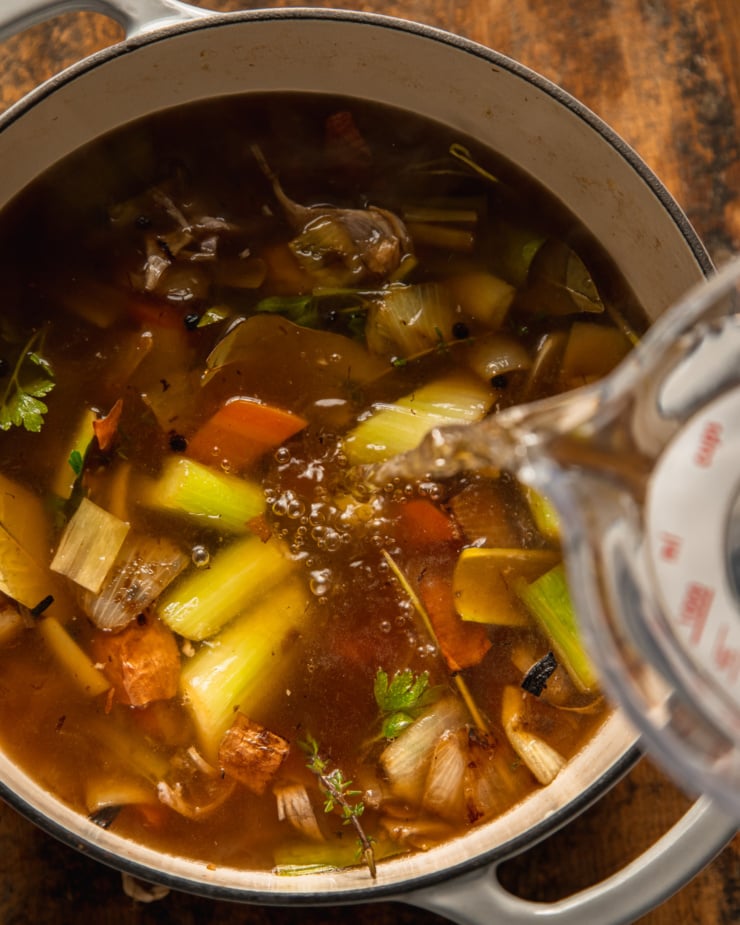 An overhead shot shows water being poured into the vegetable stock pot that is filled with vegetables and herbs.