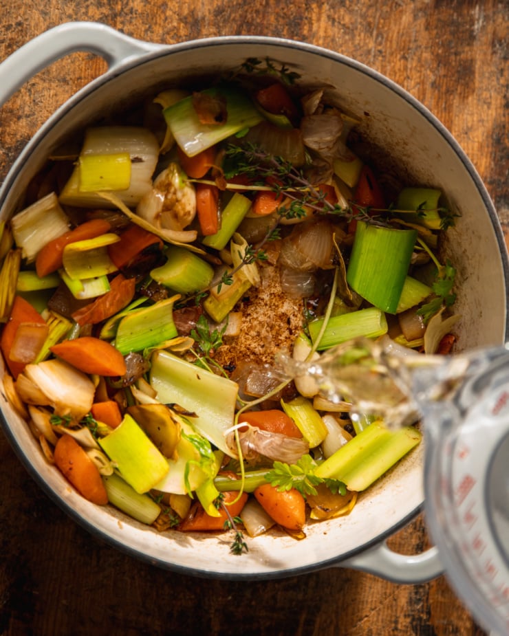 An overhead shot shows some water being poured into a pot of sautéed vegetables and herbs.