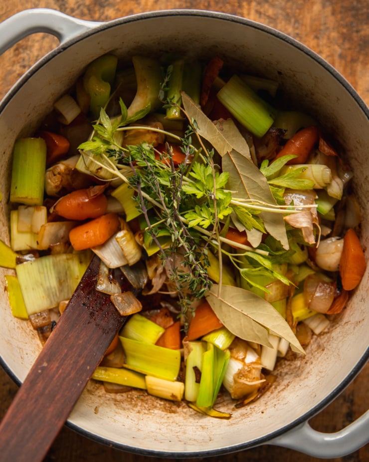 An overhead shot shows a pot filled with sautéed onions, carrots, celery, and leeks with some bay leaves and sprigs of parsley and thyme on top