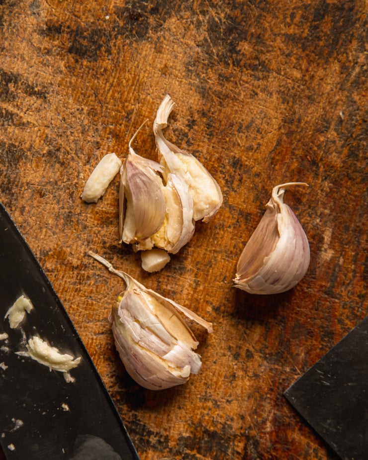 An overhead shot shows 3 garlic cloves in the process of being smashed on a rough wooden cutting board.