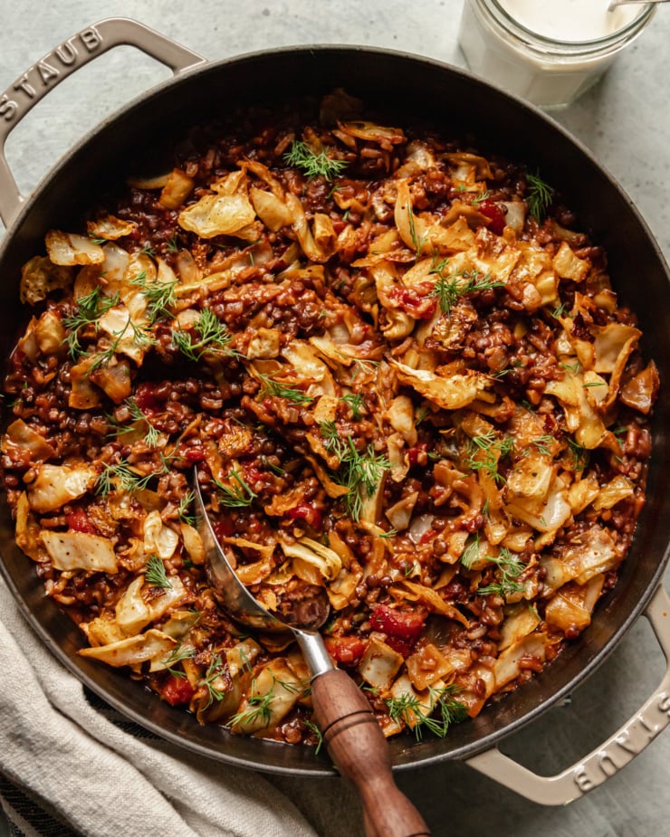 An up close, overhead shot shows a finished vegan cabbage roll skillet with french lentils, chopped tomatoes, rice, walnuts, and spices. The skillet is topped with fresh dill and a jar of vegan sour cream is seen nearby.