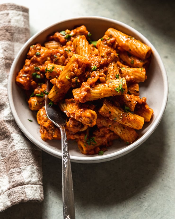 A 3/4 angle shot of a serving of lentil bolognese rigatoni in a wide, white bowl. A checkered napkin is nearby and the pasta is garnished with finely chopped parsley.
