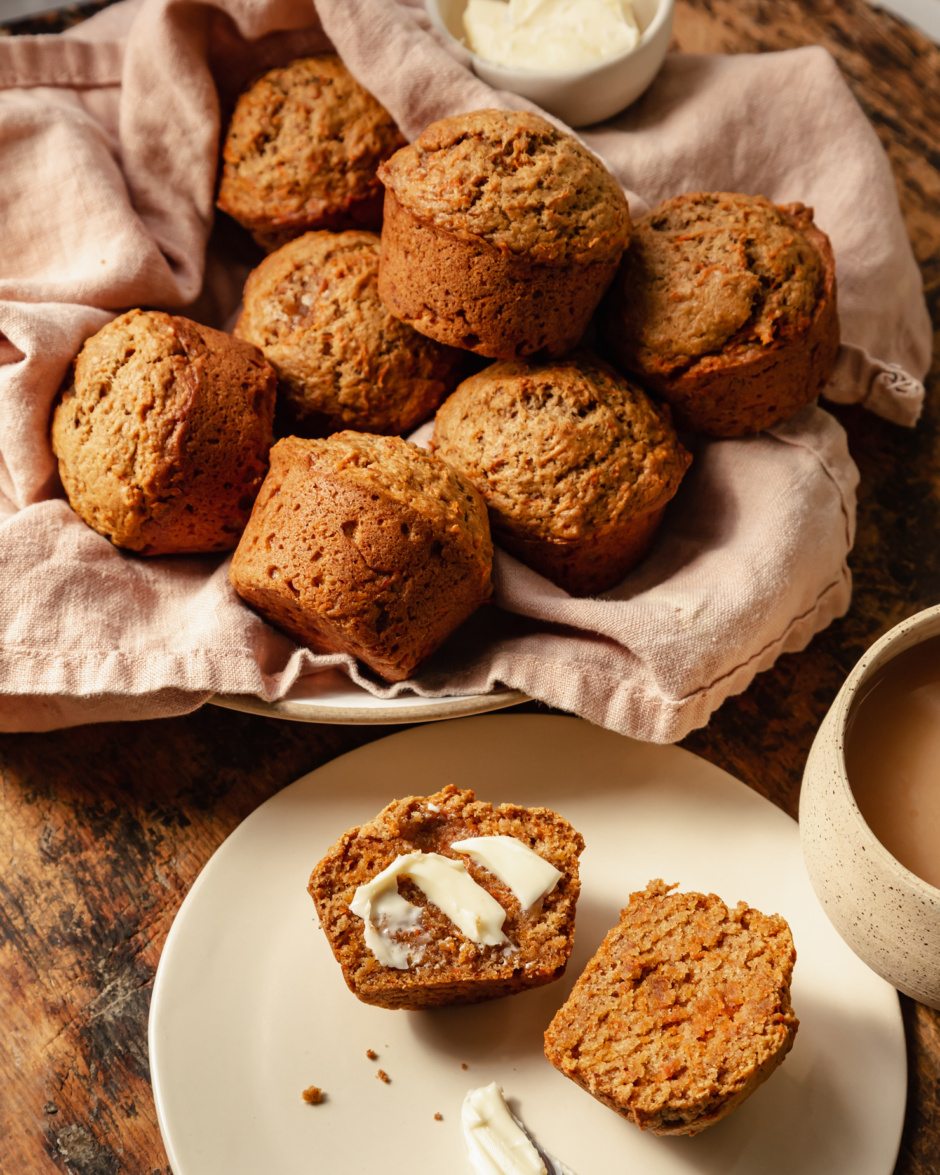 A slight 3/4 angle shot shows vegan carrot muffins in a linen-lined bowl. The muffins are golden brown with flecks of carrot, and have nicely domed tops.