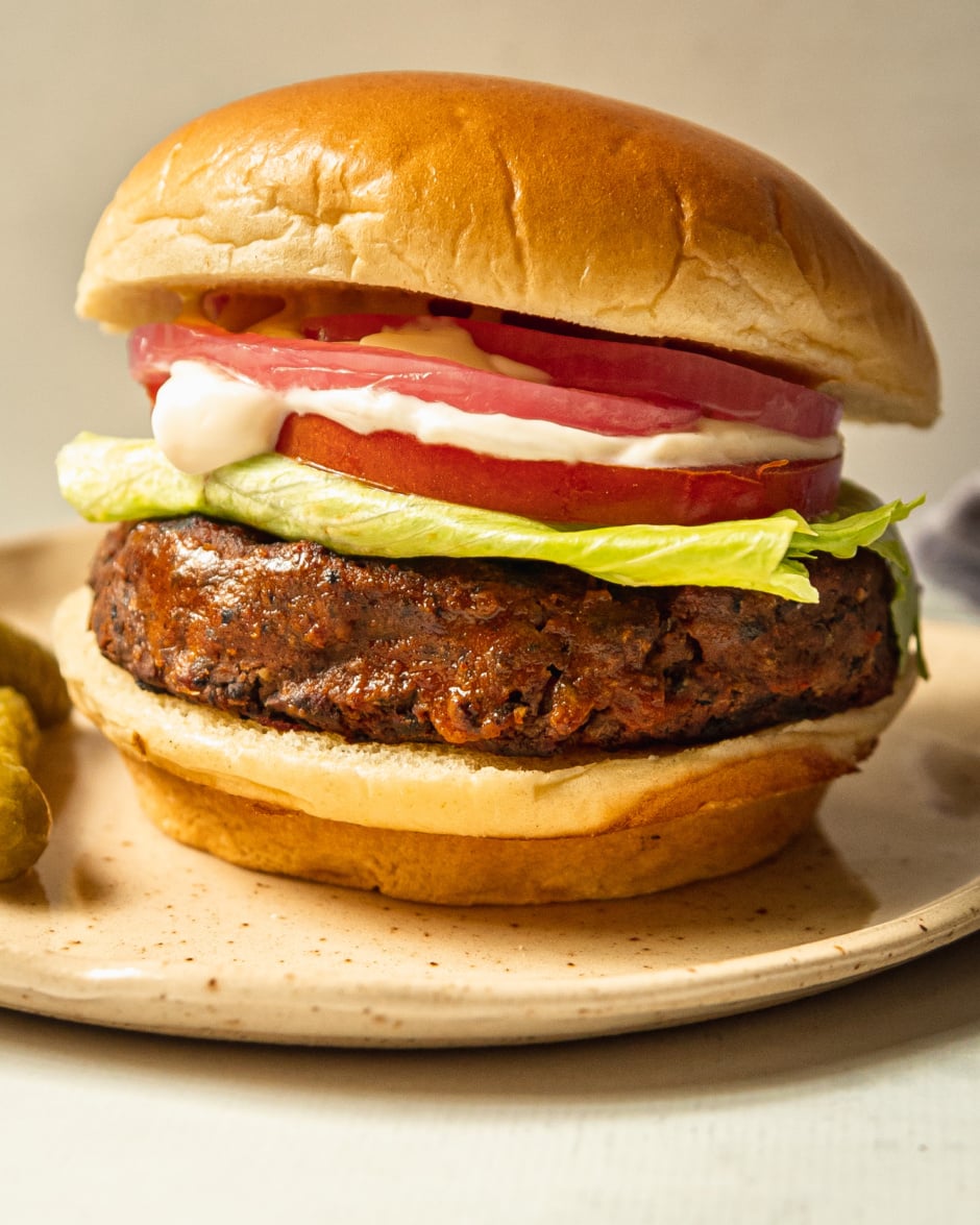 An up close, head-on shot shows a plate with a vegan black bean burger in a brioche-style bun. The burger is topped with iceberg lettuce, sliced tomato, vegan mayonnaise, mustard, and pickled red onion.