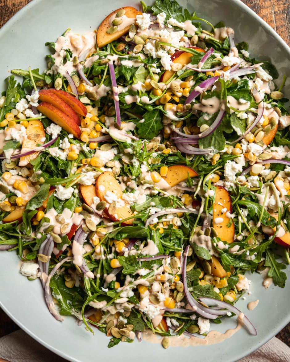 An overhead shot of a fresh summer salad featuring: cooked corn, sliced peaches, arugula, pumpkin seeds, spicy cashew dressing, and vegan feta.