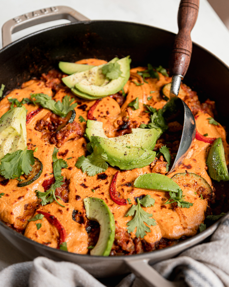 A 3/4 angle shot of lazy chopped vegan enchiladas in a skillet. A sunflower "cheese" topping blankets the top and is browned in spots. The skillet is garnished with sliced avocado and chopped cilantro.