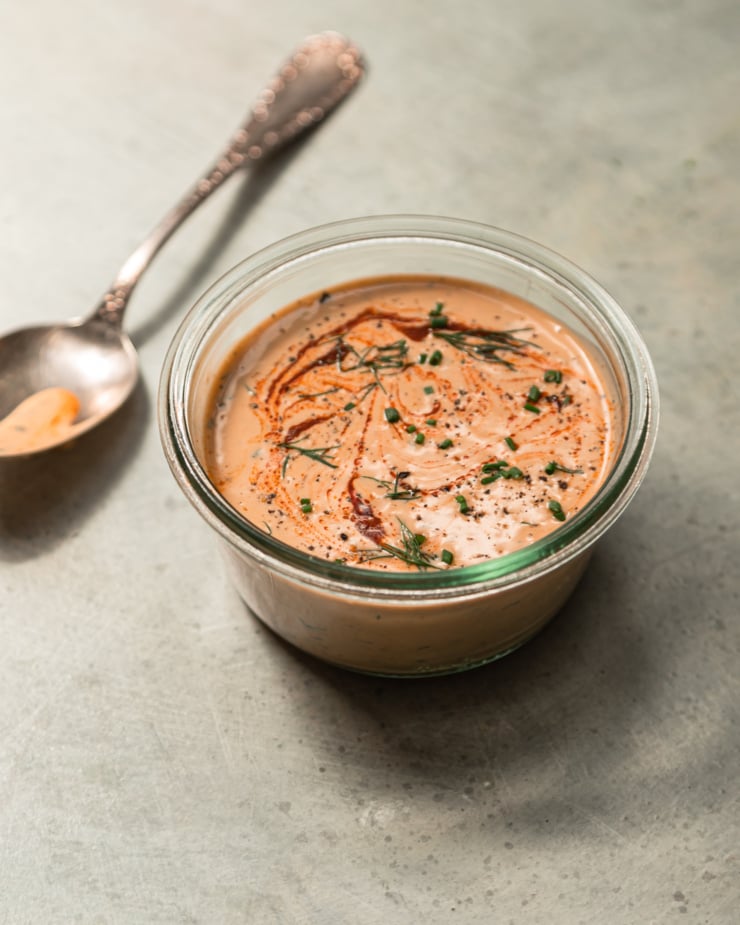 A 3/4 angle shot shows a container of spicy tahini ranch in bright natural light. The top is swirled with hot sauce and garnished with chopped herbs.