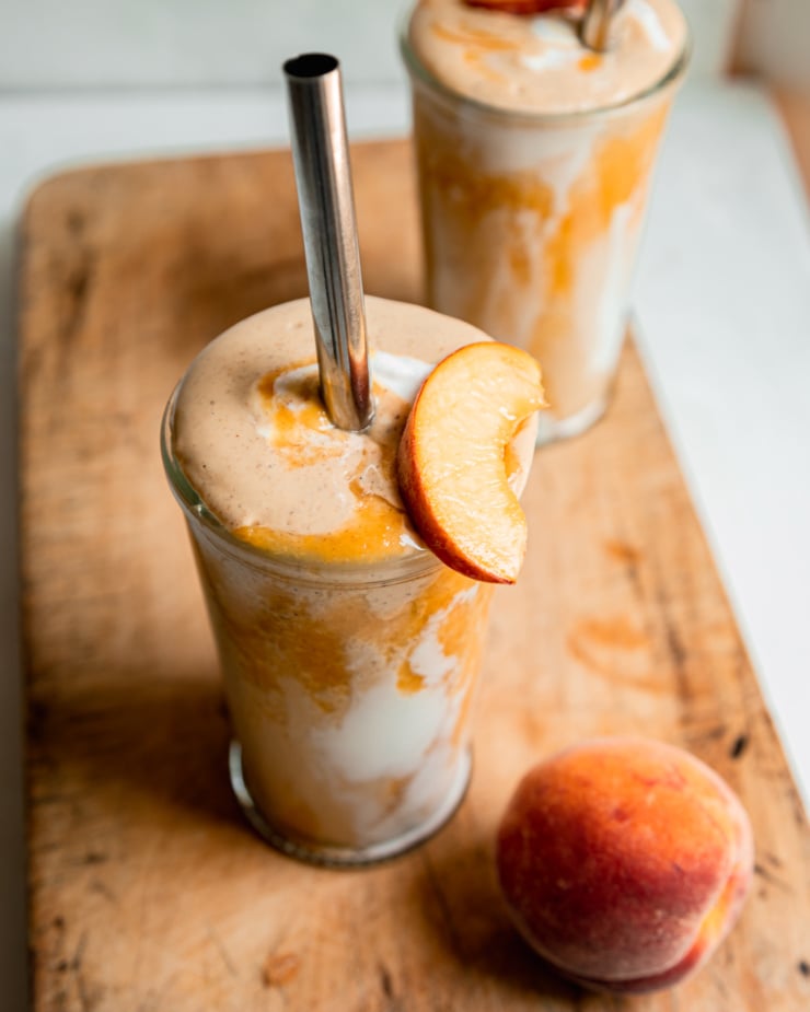 A 3/4 angle shot of a vegan peaches and cream protein smoothie in two glasses with metal straws. Peach purée and vegan yogurt create a tie dye effect on the glass. A slice of peach is perched on top of the one glass.