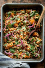 An overhead shot shows a baking dish filled with baked pearl couscous and butternut squash. The bake features chickpeas and spinach, and is topped with pickled red onions and drizzles of a creamy green basil tahini sauce. A wooden serving spoon is sticking out of the dish.