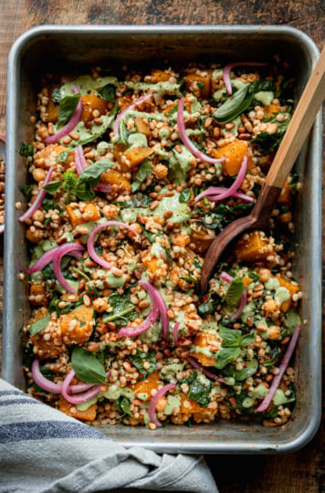 An overhead shot shows a baking dish filled with baked pearl couscous and butternut squash. The bake features chickpeas and spinach, and is topped with pickled red onions and drizzles of a creamy green basil tahini sauce. A wooden serving spoon is sticking out of the dish.