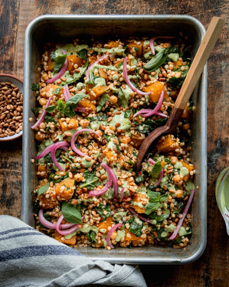 An overhead shot shows a baking dish filled with baked pearl couscous and butternut squash. The bake features chickpeas and spinach, and is topped with pickled red onions and drizzles of a creamy green basil tahini sauce. A wooden serving spoon is sticking out of the dish.