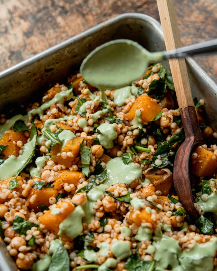 A 3/4 angle shot shows a creamy green basil tahini being drizzled from a spoon on top of baked pearl couscous with butternut squash, spinach, and chickpeas.