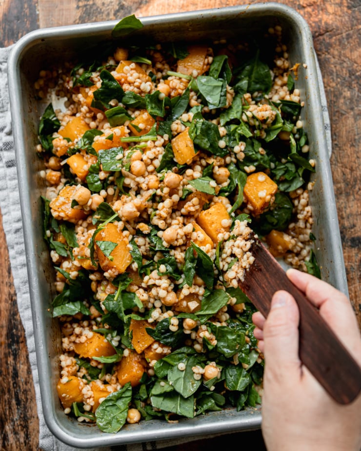 An overhead shot shows a hand using a wooden utensil to stir together some baked butternut squash, pearl couscous, chickpeas, and chopped spinach.