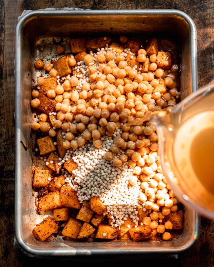 An overhead shot shows vegetable stock being poured over a pan of baked squash, chickpeas, and raw pearl couscous