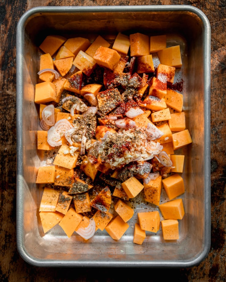 An overhead shot shows cubes of raw butternut squash topped with sliced shallots, minced garlic, smoked paprika, za'atar, salt, pepper, and olive oil; all in a metal baking dish.