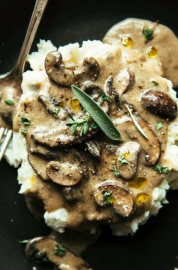 An overhead shot shows creamy mushroom gravy spooned over mashed potatoes. Fresh herbs garnish the top of this dish that is plated on a matte black plate.
