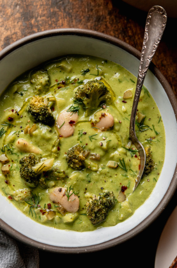 An up close overhead shot shows a bowl of broccoli dill pickle soup with white beans and chopped potatoes. A spoon is sticking out of the bowl. The soup is garnished with chili flakes, chopped fresh dill, and swirls of olive oil.