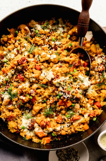 An overhead shot shows one-pot rice and veggies with chickpeas and a drizzle of creamy dilly tahini sauce on top. The dish is garnished with fresh dill, chili crisp and sesame seeds. A serving spoon is sticking out of the pot.