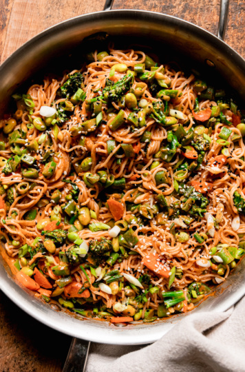 An overhead shot shows a bunch of vegan Gochujang noodles in a skillet with lots of sautéed chopped vegetables. The noodles are garnished with sliced green onions and sesame seeds.