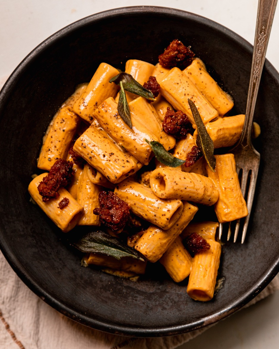 An overhead shot shows an individual serving of vegan butternut squash pasta. The sauce is very creamy and the pasta is dotted with bits of cooked veggie sausage and fried sage leaves.