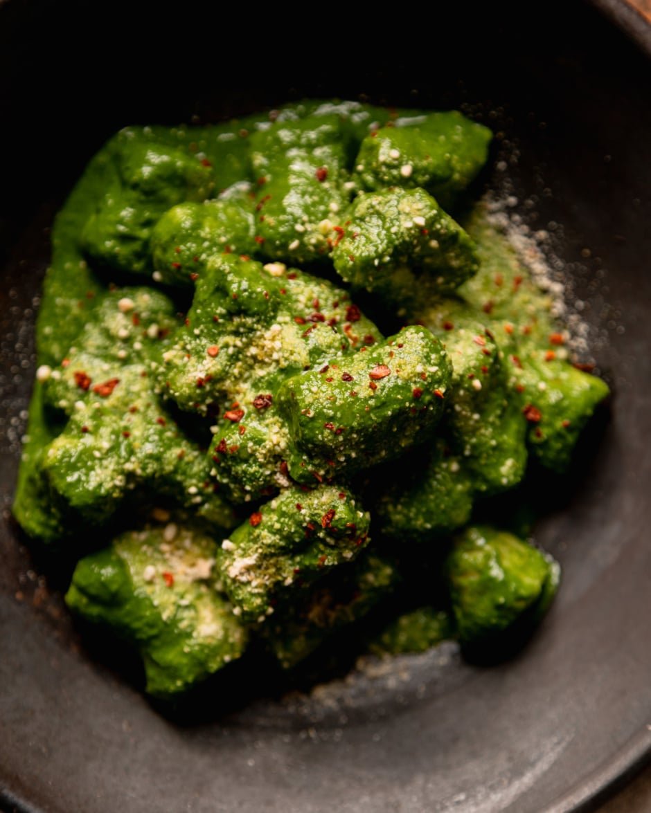 An up close, overhead shot shows a serving of homemade vegan gnocchi covered in a thick green kale sauce. The dish is garnished with vegan "parmesan" and Aleppo chili