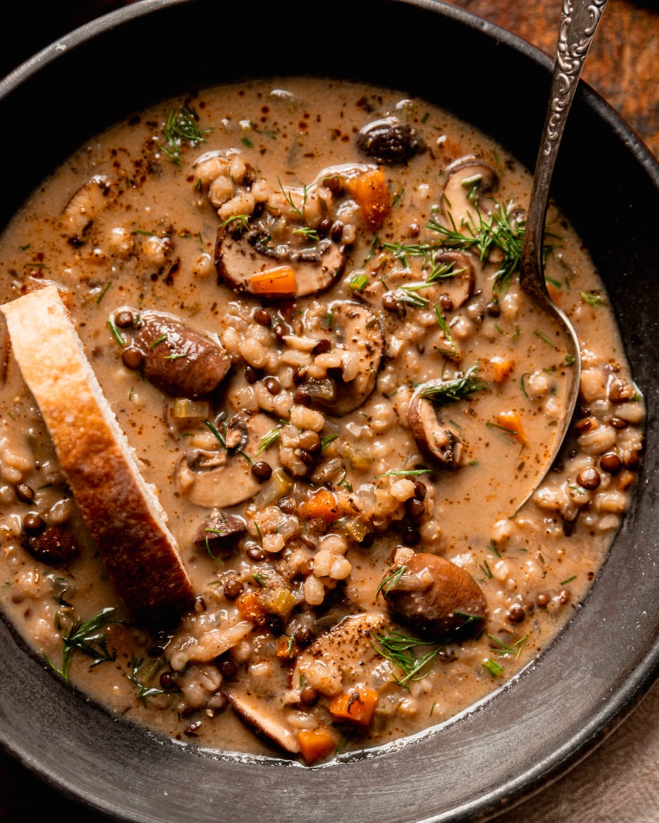An up close, overhead shot shows a creamy vegan mushroom barley soup with lentils. The soup is garnished with chopped fresh dill. A slice of bread is sticking out of the bowl of soup as well as a spoon.