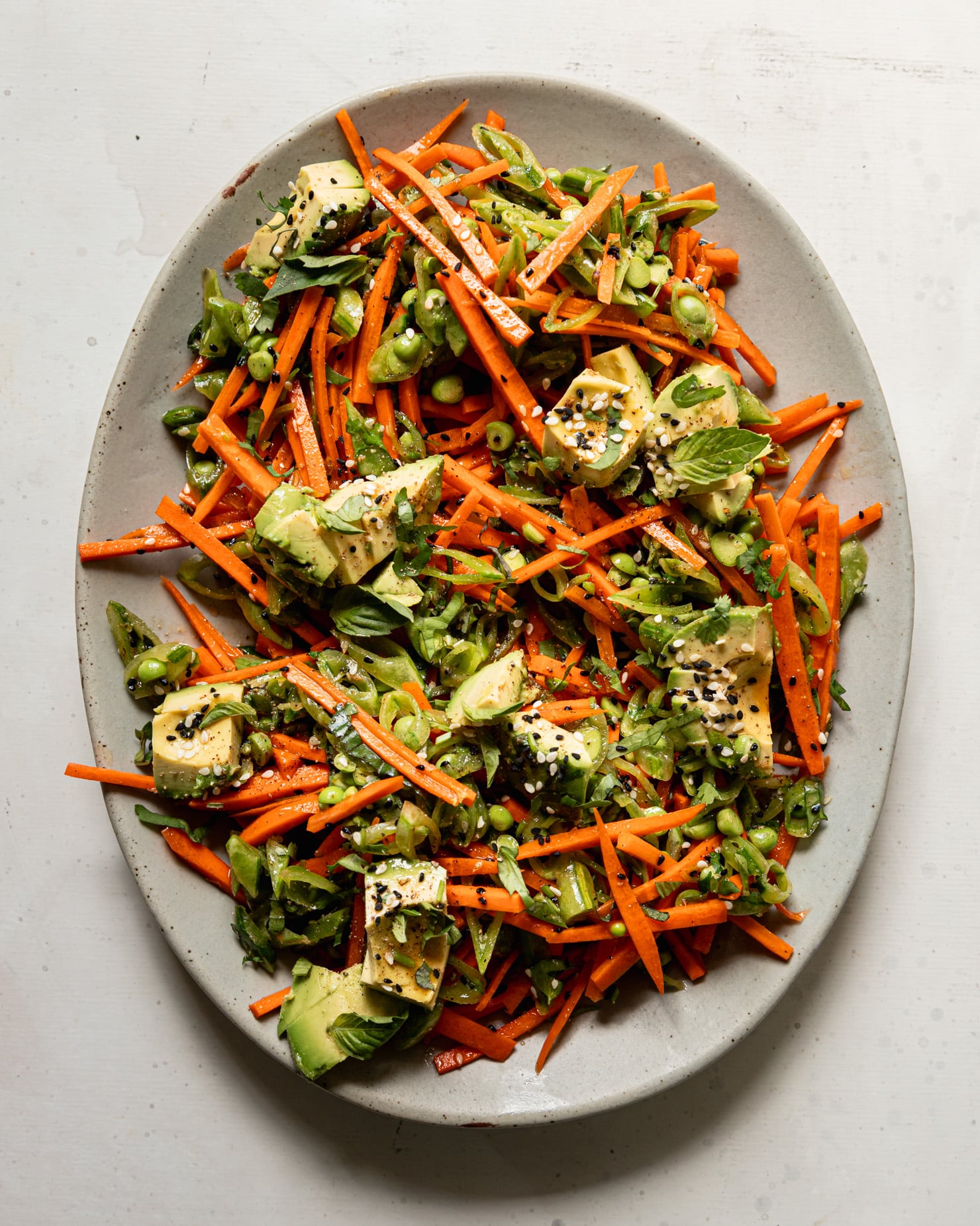 An overhead shot shows a crunchy shredded snap peas and carrots salad with chunks of avocado on top. The salad is dressed with a sesame, lime and garlic dressing. Chopped cilantro, Thai basil, and toasted sesame seeds fleck the salad throughout.