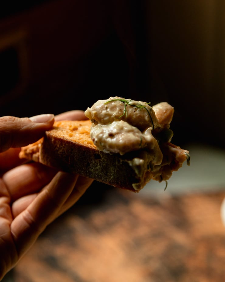 A head-on shot shows a hunk of bread with some creamy dilly beans and peas scooped on top. A hand is holding the piece of bread.