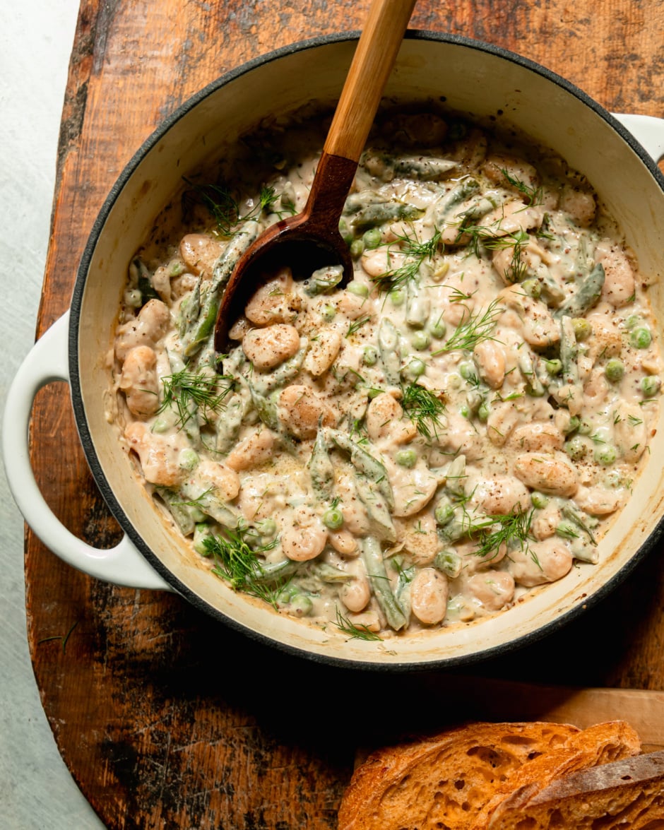 An overhead shot shows a braiser pot filled with creamy dilly beans and peas. A serving spoon sticks out of the pot and some bread is seen to the side.