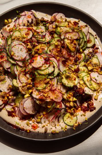 A brightly lit, overhead shot shows a shaved cucumber radish salad on a moat of garlic lime tahini. The salad is garnished with finely chopped pistachios and chili crisp.