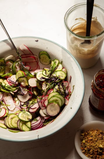 A 3/4 angle shot shows a shaved radish and cucumber salad in a bowl, a tahini-based sauce, a jar of chili crisp, and finely chopped pistachios.