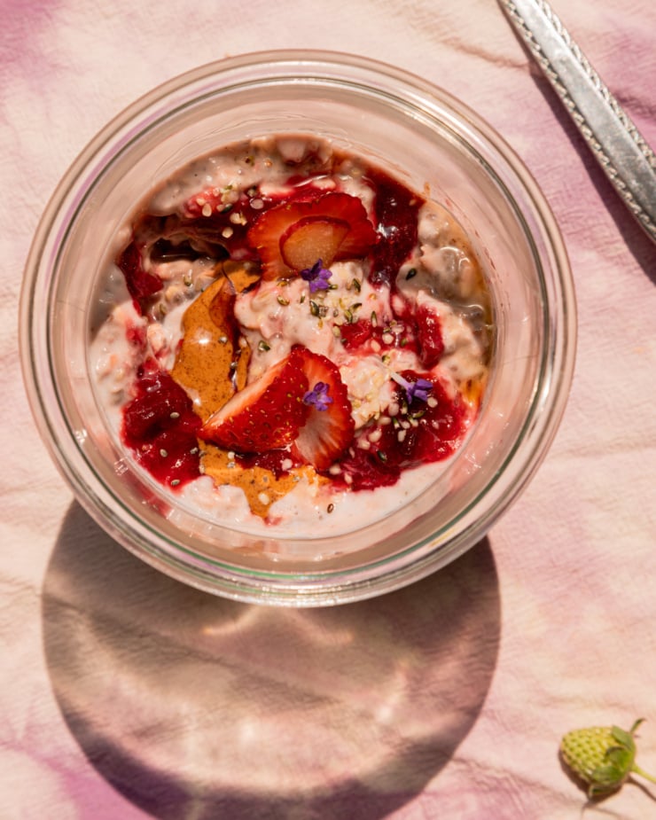 An up close, overhead shot shows a serving of strawberry rhubarb overnight oats in a glass jar. The top is garnished with fresh strawberries, almond butter, compote, hemp seeds, and lavender flowers. A teeny green strawberry on the stem is seen in the bottom corner.