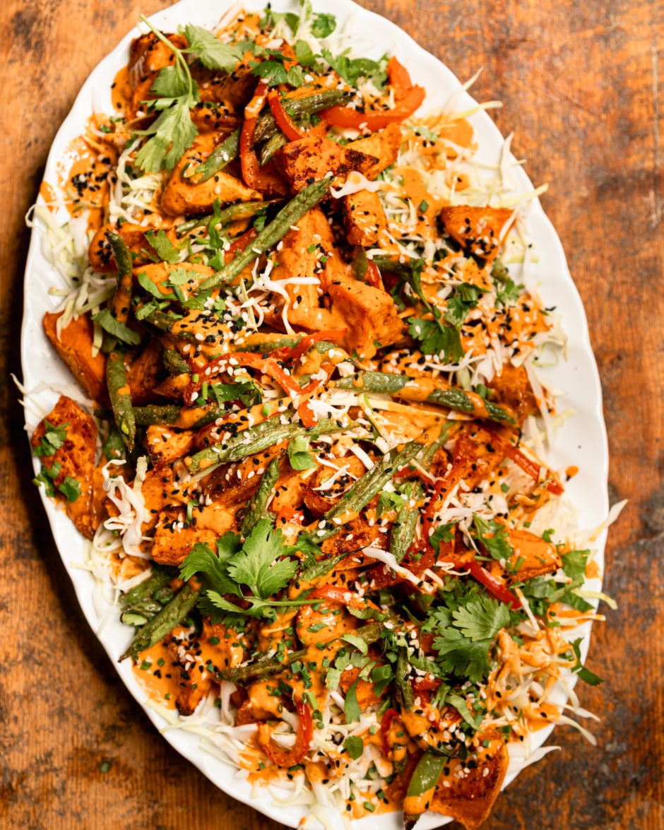 An overhead shot shows a Gochujang peanut sweet potato salad with green beans, red bell peppers, shredded cabbage, and chopped herbs. The roasted vegetables are piled on top of the cabbage on an oval platter and topped with peanut Gochujang dressing and sesame seeds.
