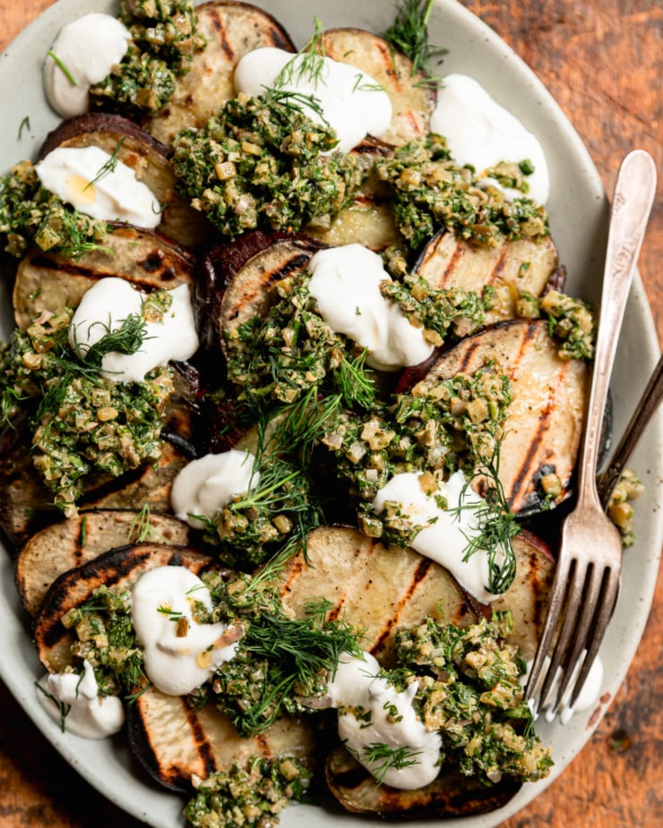 A close-up overhead shot shows a pile of roasted Japanese sweet potato chips topped with a ball of dill pickle salsa verde, vegetarian sour cream and fresh dill.