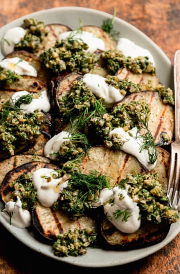 A 3/4 angle shot shows a platter of grilled Japanese sweet potato slices topped with dollops of dill pickle salsa verde, dollops of vegan sour cream, and fresh dill.