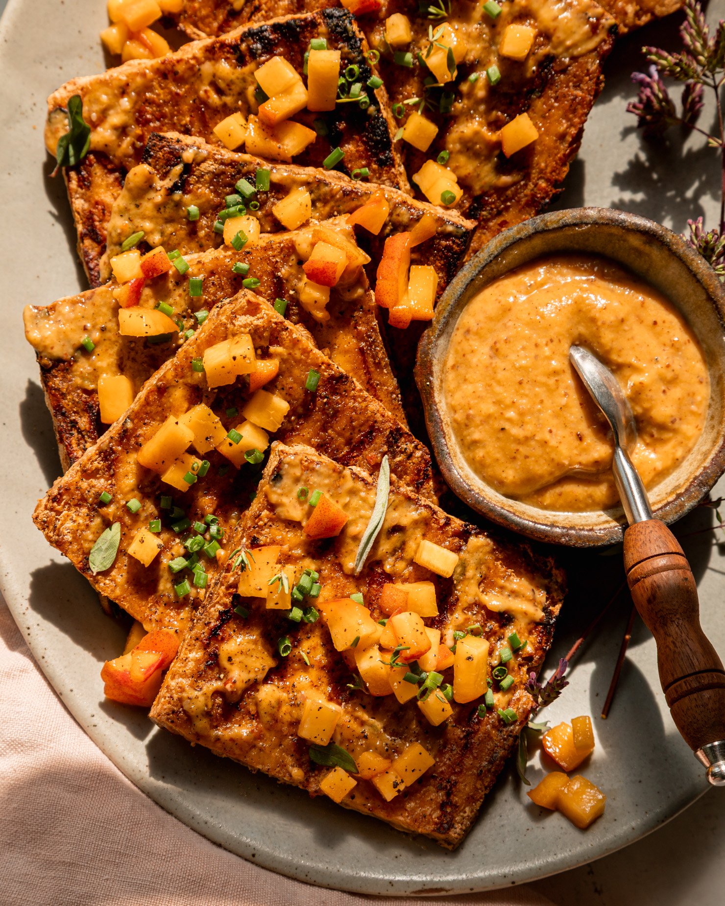 A close-up overhead shot shows a plate with baked tofu slices coated with peaches, almond butter and a flavour glaze. Tofu is decorated with small peaches and chopped herbs. A small bowl of sauce can be seen on the side of the plate. This photo was taken in direct sunlight with some harsh shadows.