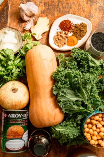 An overhead shot shows ingredients needed for a butternut squash stew with chickpeas and kale. All ingredients are on a rough wood board.