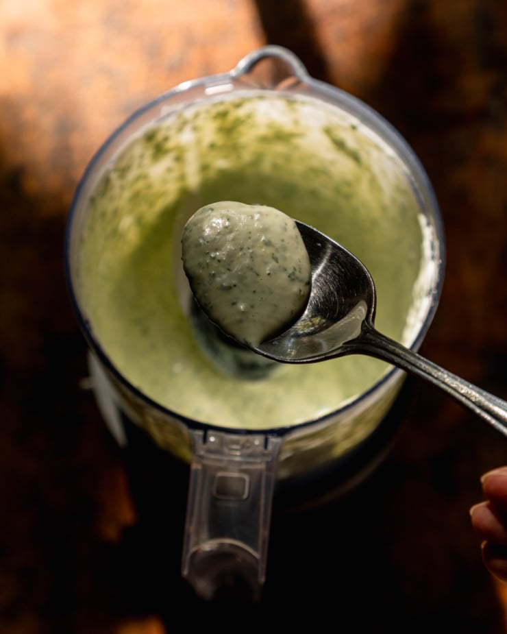 The overhead shot shows a spoonful of creamy green coriander sauce hovering over a mini food processor filled with the same sauce.
