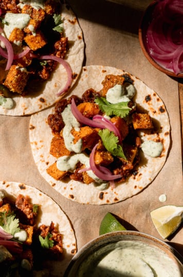 A overhead image shows 3 vegan butternut squash tacos with creamy cilantro sauce drizzled on top. The tacos also feature a smashed chipotle pinto bean base, chopped cilantro, and pickled red onions. Lime wedges and a bowl of the sauce are seen nearby.