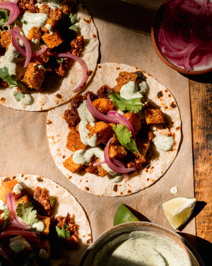 The top picture shows 3 vegan butternut squash tacos topped with creamy cilantro sauce. The tacos are also equipped with crushed Chipotle Pinto bean base, chopped cilantro and pickled red onions. See lime wedges and a bowl of sauce nearby.