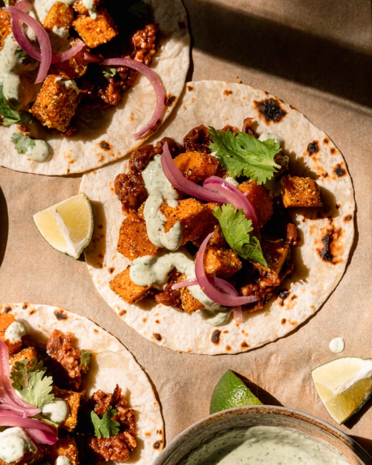 Close-up overhead image shows 3 vegan butternut squash tacos with creamy cilantro sauce drizzled over it. The tacos are also equipped with crushed Chipotle Pinto bean base, chopped cilantro and pickled red onions. Lime wedges are seen nearby.